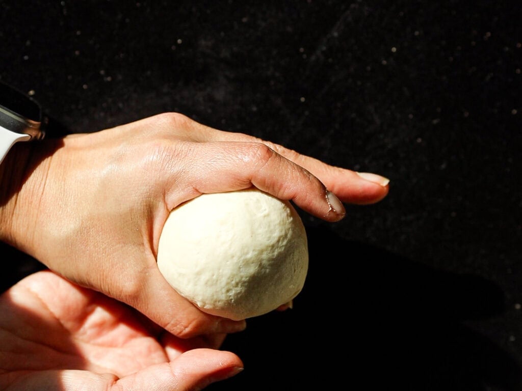 A person shaping dough into a round ball with their hands against a dark background, indicating a baking or cooking process.